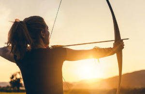 Woman with a bow and arrow aiming, sunlight behind